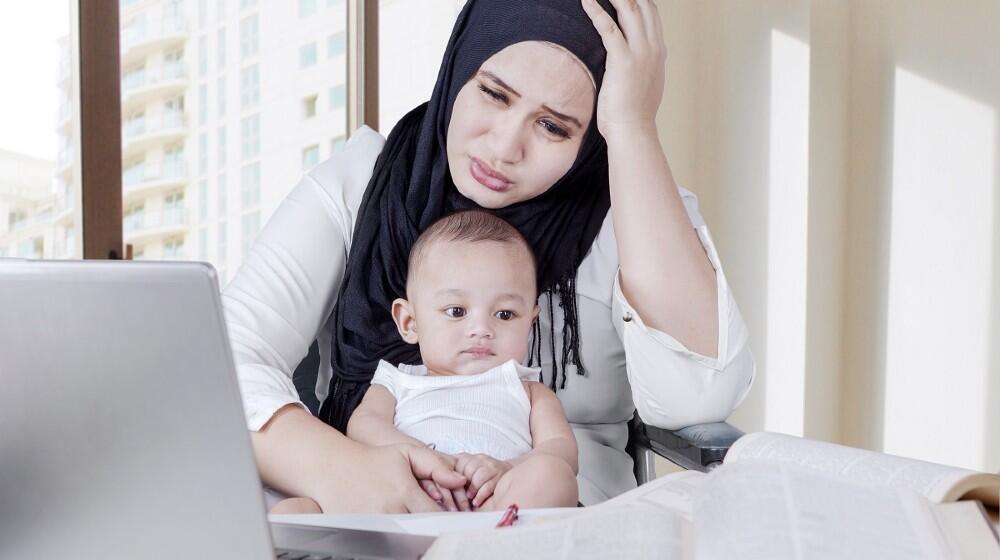 A mother looking stressed while on a laptop while holding a baby.